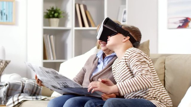 Medium shot of teenage boy and his grandmother sitting on sofa in living room. Woman reading newspaper while boy enjoying VR goggles