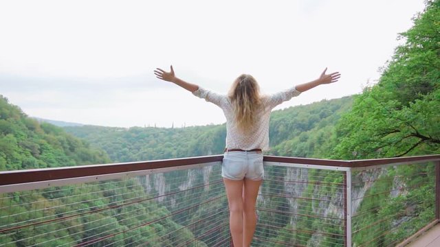 Happy woman blond hair in casual clothes stands back, young girl  hands raised to sky, tourist cheerfull and joyfully enjoying summer green nature mountains Georgia, Martvili canyon, observation point