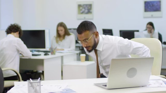 Medium shot of Arab businessman leaning on desk with his hands and doing push-ups while his colleagues working on background