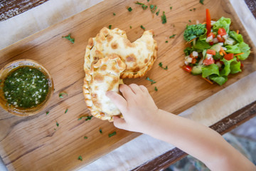 Child eating baked stuffed pastry, top view at the hand taking empanadas from wooden plate