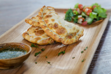 Empanadas, baked stuffed pastry served on a wooden plate with fresh green salad and parsley dip