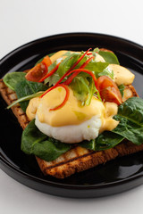 Side close view of grilled italian sandwich on wheat toast with greens, benedict egg, chili pepper and cherry tomatoes, served on black dishware and isolated on white