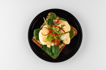 Top view of grilled italian sandwich on wheat toast with greens, benedict egg, chili pepper and cherry tomatoes, served on black dishware and isolated on white