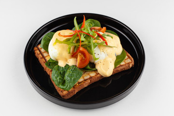 Side close view of grilled italian sandwich on wheat toast with greens, benedict egg, chili pepper and cherry tomatoes, served on black dishware and isolated on white