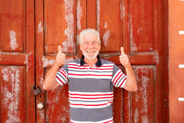 People senior with white beard and hair standing behind an old red door closed by lock. spanish home with red windows and doors made by wood. Rustic style. Sunlight in summer time. Old man smiles