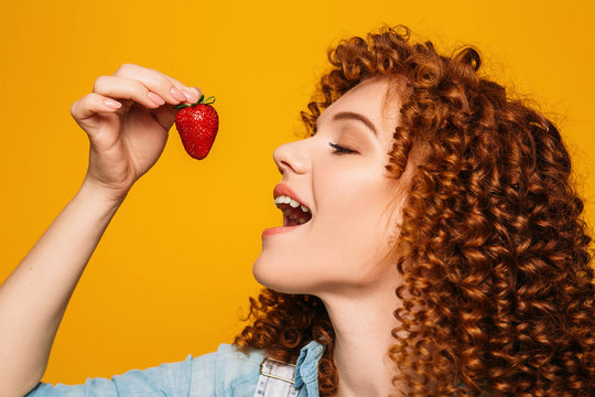 curly red-haired woman with open mouth wants to eat delicious strawberries on a yellow background.