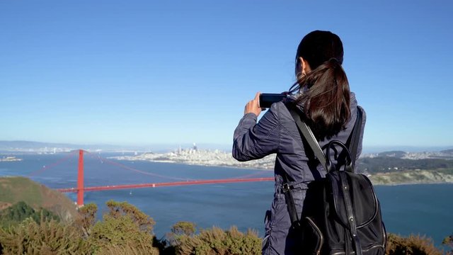 nature lover female asian travel in san francisco usa. back view young chinese woman take picture of beautiful amazing landscape golden gate bridge with blue sea ocean sky on mobile phone outdoor.
