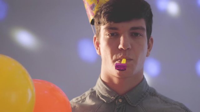 Portrait of young cute man in birthday hat with noisemaker in mouth looking in the camera holding balloons. Bright lights of different colors on his face and on the wall. The guy at the party
