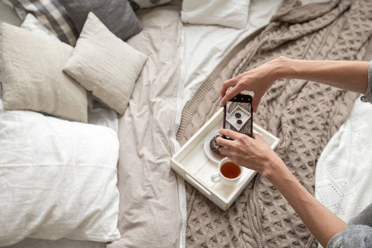 From above shot of woman sitting on bed and taking picture of tea and doughnut on tray with smartphone