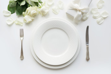 White table setting with bouquet rose, gift, dishware, silverware on holiday white table. Top view. Romantic and wedding anniversary.