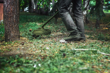 lawnmower or lawn keeper in dirty working boots and special clothes works in the park and cuts the grass by garden trimmer; scraps of foliage fly apart