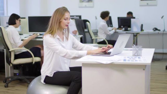 Lockdown of blonde businesswoman sitting on fitball at desktop and using laptop while her colleagues working on background