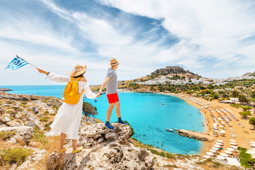 A couple of friends in love with the Greek flag admire the grandiose view of the sea Bay and the beach near the ancient city of Lindos. Honeymoon and travel © EdNurg