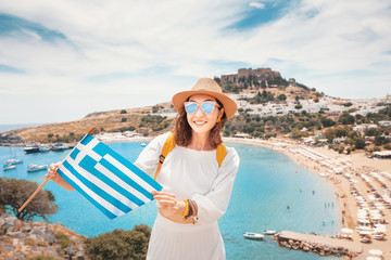 Elegant asian woman in white dress posing with greek flag in front of the famous Lindos town on Rhodes island. Vacation and travel in Greece
