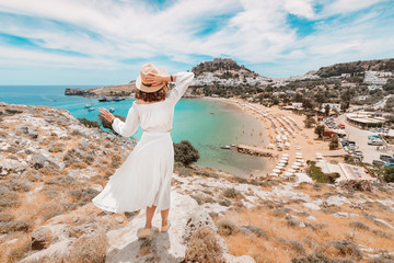 Happy asian woman travels in Greece. Having fun at the stunning view on sea resort and old town of Lindos on the mountain - obrazy, fototapety, plakaty