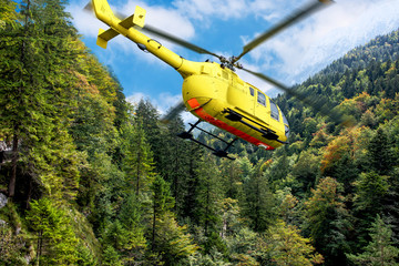 Fototapeta na wymiar Yellow helicopter on background of high Alps mountains wth green forest under blue cloudy sky.