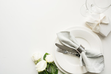 White table setting with bouquet rose, gift, dishware, silverware on holiday white table. View from...