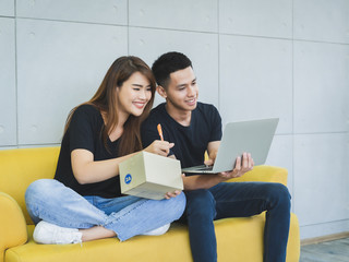Young happy asian business couple is work together by using laptop, smartphone or tablet with a parcel box packaging at their startup home office, SME online business seller and delivery concept