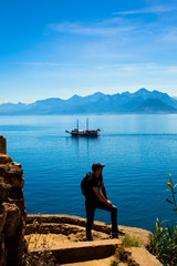 Fototapeta na wymiar Young man stands on a rock on the background of a beautiful view of the Mediterranean Sea, the mountains and the ship. Turkey, Antalya.