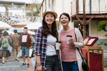 two asian girls friends standing on street in holidays event fair outdoor in village on sunny day. young women smiling laughing chatting looking with funny thing creative market in thailand bangkok