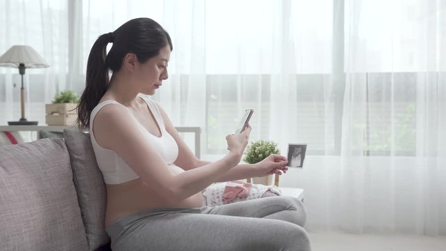 pregnant woman holding ultrasound scan and taking picture by cellphone. future mom with bare tummy relax on sofa using smart phone. young wife motherhood sending photo of baby sharing to husband