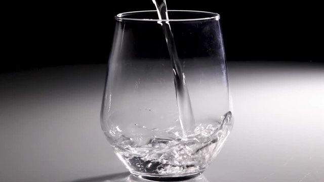 Slow-Motion shot of water being poured into a clear glass tumbler with a dark background.
