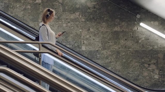 Young beautiful girl goes down the escalator in the subway and looks into the smartphone. A student with headphones goes to the university. Listening to music on headphones in the smartphone.
