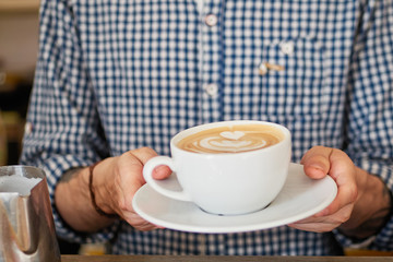 Close up of barista man hands serving freshly brewed cappucino coffee while standing in coffee shop. Focus on male hands holding a big white ceramic cup of coffee with copyspace