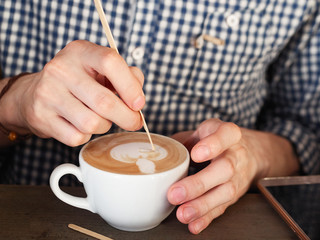 Close up shot of barista in white blue checkered shirt hands making latte art at the freshly brewed cappuccino in big white ceramic mug with a wooden coffee stick in cozy craft cafe