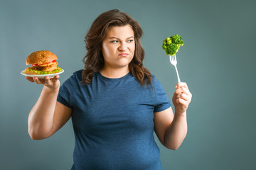 Indignant plus size woman with healthy and unhealthy food on grey background. Concept of weight loss