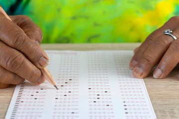 Close-up image of elderly asian woman hands doing exams by using pencil selected multiple choice on standardized test form with answers bubble on wood table. Education and Lifelong learning concept.