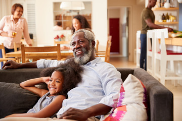 Grandfather With Granddaughter Sitting On Sofa At Home Watching Movie With Family In Background