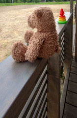 A brown toy bear sits on a wooden railing, next to a children's toy pyramid on the terrace of a country house. Vertical orientation, space for text.