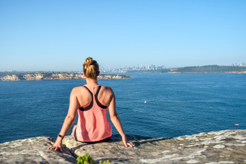 Morning view of a young female hiker sitting on a rock at North Head, a headland in Manly and part of Sydney Harbour National Park in Sydney, New South Wales, Australia. Sydney skyline in background.