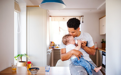 A father bottle feeding a small toddler son indoors at home.