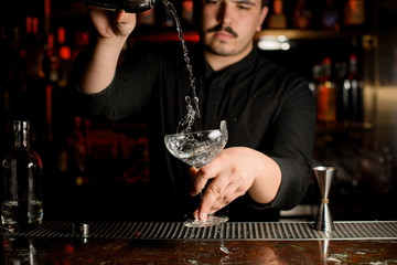 Male bartender pouring a transparent alcohol into the cocktail glass from the steel shaker