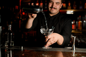 Smiling bartender pouring a transparent alcohol into the cocktail glass from the steel shaker
