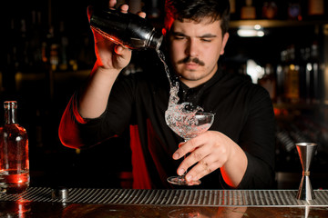 Professional bartender pouring a transparent alcoholic drink into the cocktail glass from the steel...