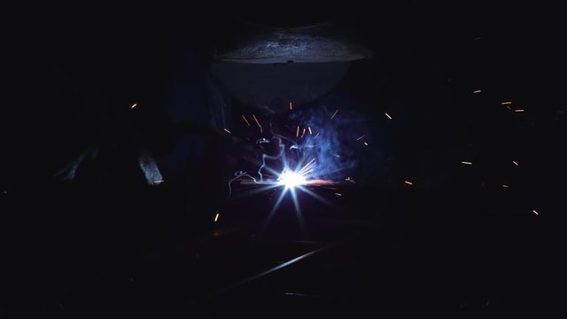A blacksmith with a welding mask and welders leathers, is welding metal products in the garage, fire sparks fly to the sides. Dark mood. Slow Motion shot with Red Monstro 8K.