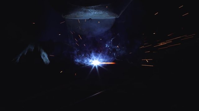 A blacksmith with a welding mask and welders leathers, is welding metal products in the garage, fire sparks fly to the sides. Dark mood. Slow Motion shot with Red Monstro 8K.