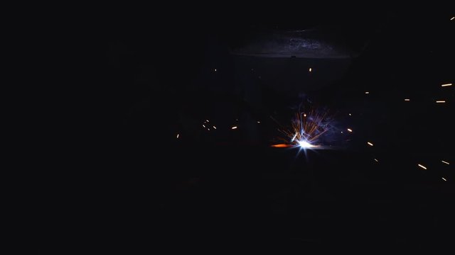 A blacksmith with a welding mask and welders leathers, is welding metal products in the garage, fire sparks fly to the sides. Dark mood. Slow Motion shot with Red Monstro 8K.