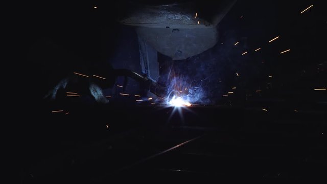 A blacksmith with a welding mask and welders leathers, is welding metal products in the garage, fire sparks fly to the sides. Dark mood. Slow Motion shot with Red Monstro 8K.