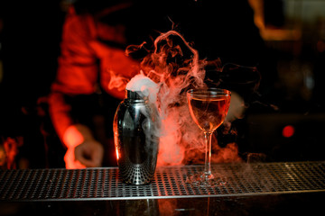 Bartender standing in the foreground of cocktail in the glass and smoky steel shaker