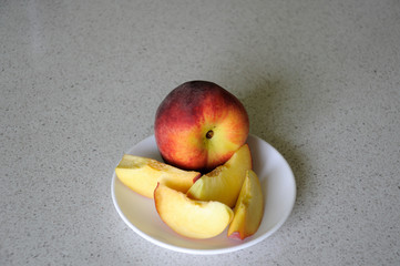 Whole red peach and a few sliced peach on a white plate on the table on a light background.