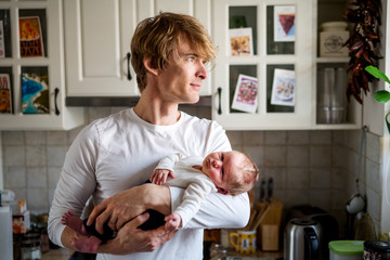 Young father holding a newborn baby in kitchen at home.