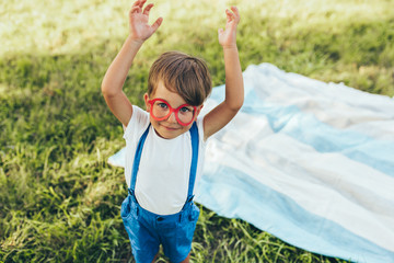 Happy little boy wearing blue shorts and red glasses playing at nature background. Cheerful child enjoying summertime in the park. Kid having fun in the forest on sunlight outdoors. Happy childhood.