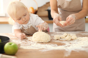 Little girl and her blonde mom in beige aprons playing and laughing while kneading the dough in kitchen. Homemade pastry for bread, pizza or bake cookies. Family fun and cooking concept