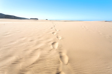 Fototapeta na wymiar Sand dunes with human footprints near seaside. Hot sunny day in deserted place
