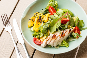 Salad with grilled chicken, mango, lettuce, avocado, tomatoes, arugula, cheese sause on a white plate on wooden background