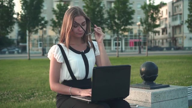 Caucasian young business woman in white shirt and glasses sits on a bench in the street and works behind a laptop. Close-up, the girl puts the glasses on her eyes and continues to work.
